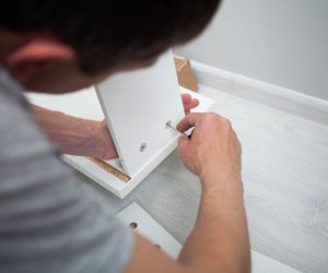 Close up of man hand, furniture assembler make flat pack white furniture assembly service at home. Person assembling white furniture, using an Allen wrench to tighten a screw into a panel on a light-colored floor.