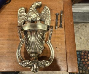Brass door knocker with eagle design placed on a wooden surface next to two screws.
