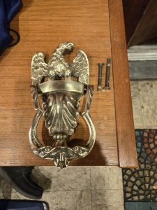 Brass door knocker with eagle design placed on a wooden surface next to two screws.
