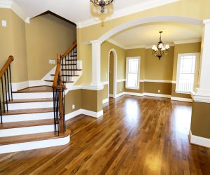 Empty, well-lit room with wooden floors, arched entryways, staircase, and beige walls with white trim and columns.