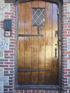 Wooden arched door with metal hinges, a small diamond window, handle, keypad lock, and a mailbox on a brick wall.