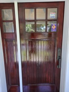 A red wooden front door with glass panels, a mail slot, two stickers, and a metal handle, reflecting outdoor greenery.