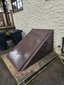 Brown metal cellar doors set in concrete at the base of a stucco house, next to garbage bins and a water spigot.
